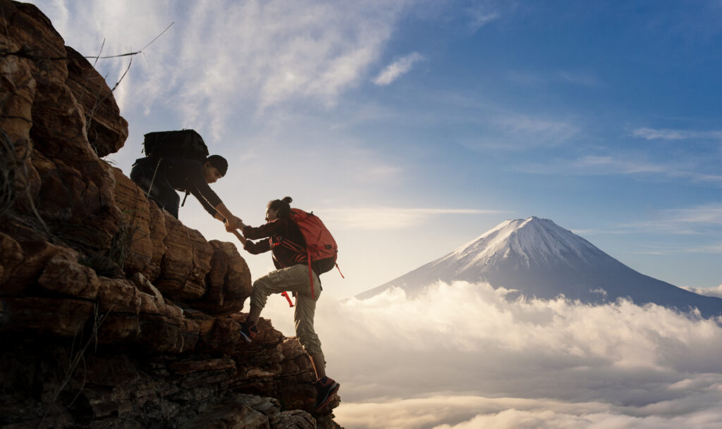 Two hikers, equipped with backpacks, help each other climb a rocky ridge above a sea of clouds, with Mount Fuji rising majestically in the background under a bright blue sky