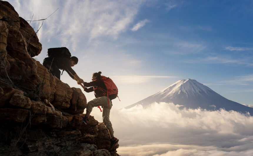 Two hikers, equipped with backpacks, help each other climb a rocky ridge above a sea of clouds, with Mount Fuji rising majestically in the background under a bright blue sky