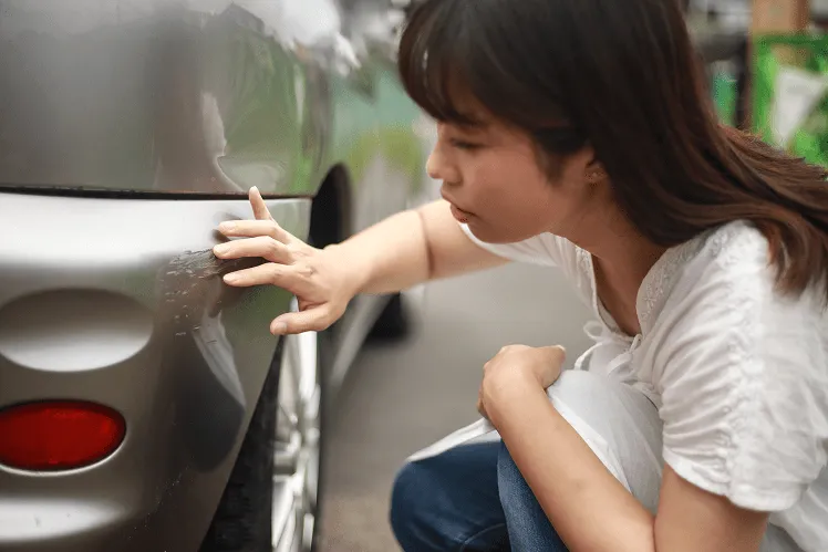 A girl closely examining a scratch on the side of her rental car, reflecting her concern after a minor accident during her trip