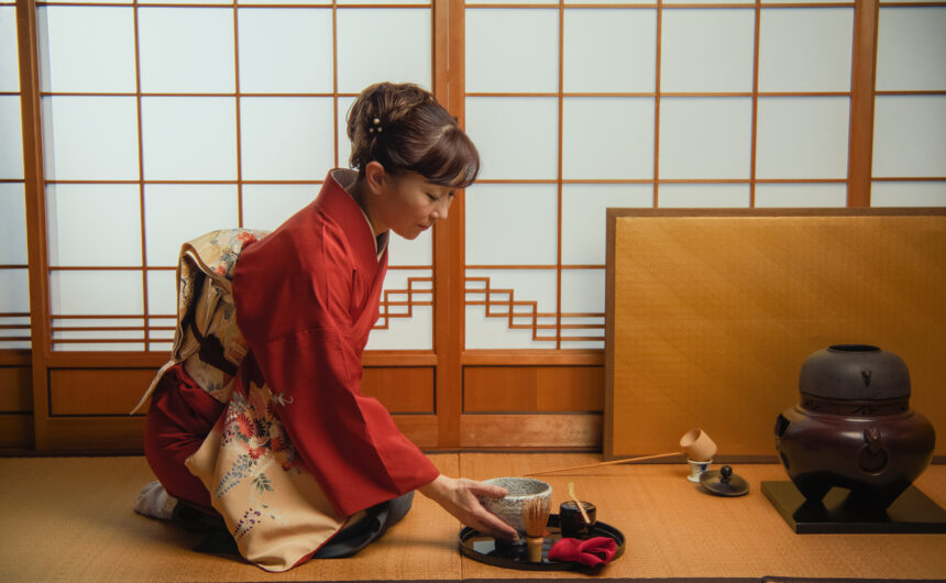 Japanese woman in a traditional red kimono with floral ornaments sits on a tatami floor, carefully preparing and sipping matcha green tea from a bowl
