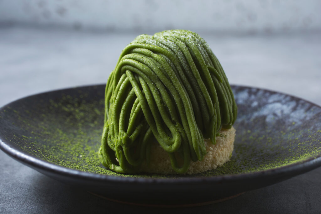 A black plate holds a beautifully crafted matcha Mont Blanc cake, its delicate strands of green tea cream artistically swirled on top