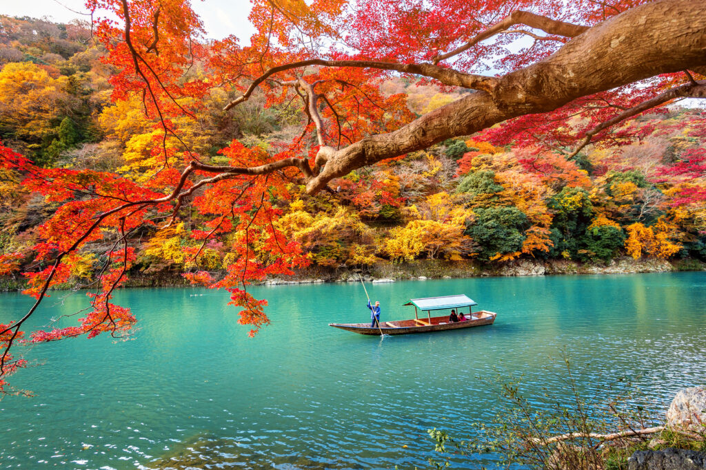 A boatman punts a wooden boat along the crystal-clear emerald river of Arashiyama, surrounded by vibrant autumn foliage in Kyoto, Japan