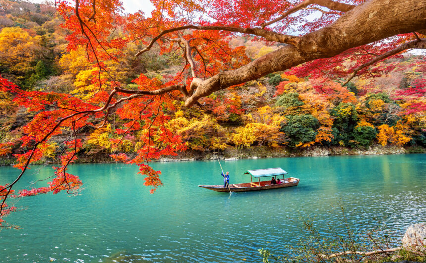 A boatman punts a wooden boat along the crystal-clear emerald river of Arashiyama, surrounded by vibrant autumn foliage in Kyoto, Japan