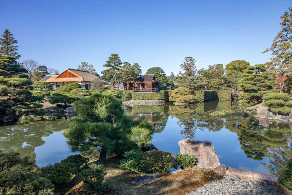 Serene view of Katsura Imperial Villa in Kyoto, Japan, nestled amid lush green trees with a tranquil pond nestled between them, all bathed in bright sunlight under a clear blue sky