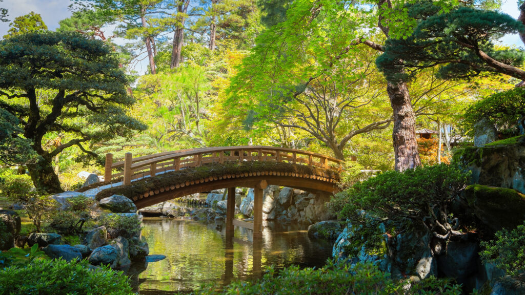 View of the Kyoto Imperial Palace with the Gonaitei garden in Kyoto, Japan, on a bright summer day. The scene features lush green trees, sunlight reflecting off the water beneath a graceful bridge, capturing the serene beauty of traditional Japanese landscaping within the palace grounds
