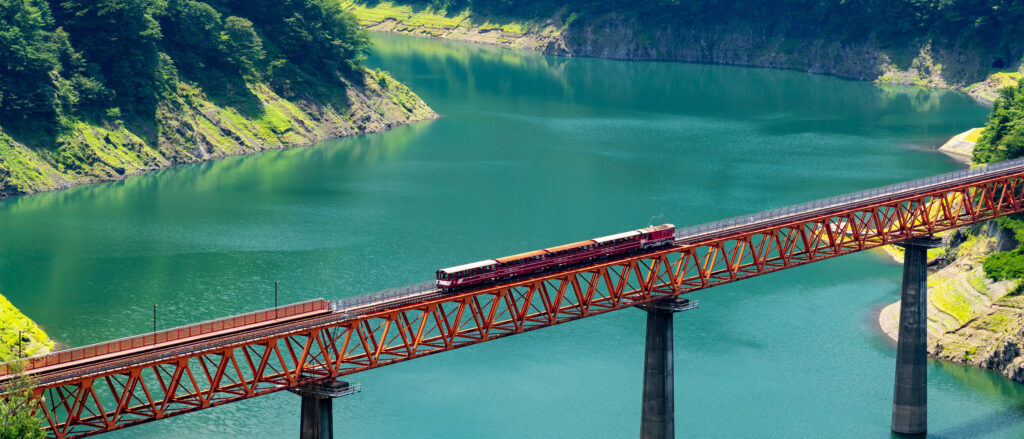 On a bright sunny day, the scene captures the harmonious blend of human engineering and nature, with the vibrant color of the train, striking red bridge, and the clear waters below