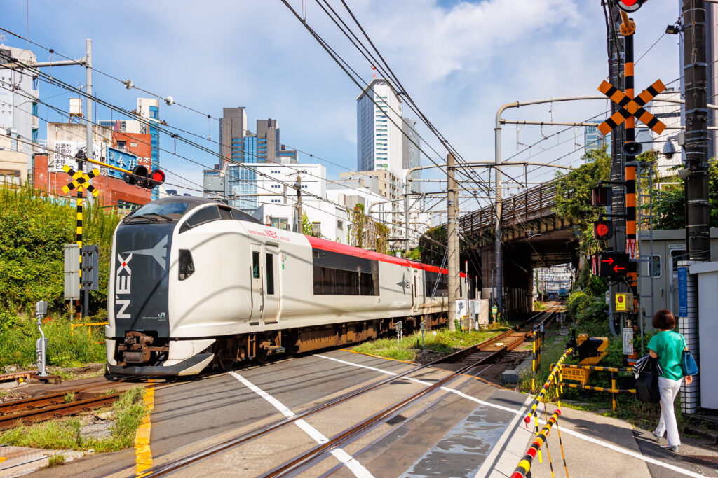 A Narita Express (N’EX) train is seen running through the heart of the city, surrounded by tall modern buildings. A woman stands patiently at the pedestrian crossing, waiting for the train to pass before she can safely cross the road