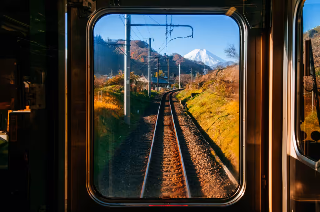 A view from inside a train frames a single railway track curving gently into the distance, leading the eye straight toward a snow-capped mountain under a clear blue sky
