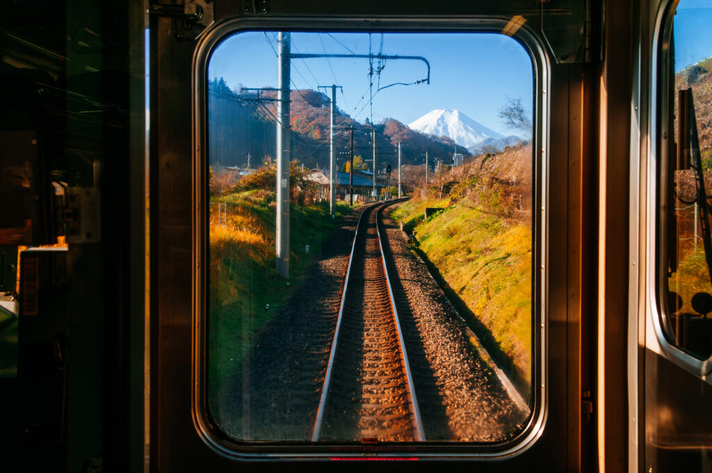 A view from inside a train frames a single railway track curving gently into the distance, leading the eye straight toward a snow-capped mountain under a clear blue sky