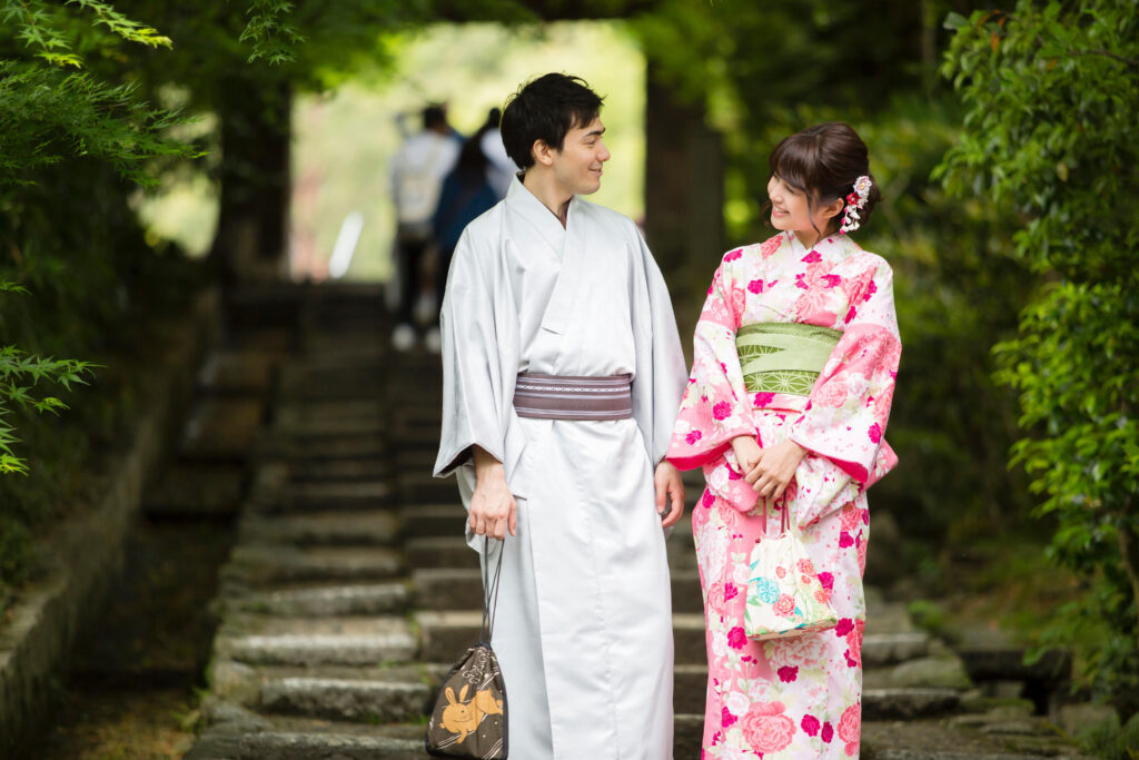 Kimono Experience Kyoto. A smiling couple stands side by side, looking radiant in their traditional kimonos. The man wears a crisp white kimono that gives him a clean and elegant look, while the woman is dressed in a soft pink kimono decorated with delicate patterns