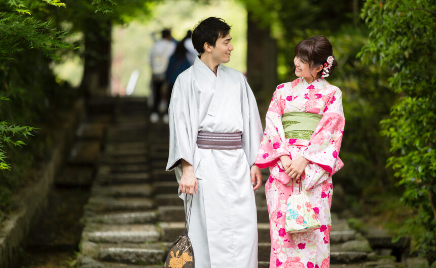 Kimono Experience Kyoto. A smiling couple stands side by side, looking radiant in their traditional kimonos. The man wears a crisp white kimono that gives him a clean and elegant look, while the woman is dressed in a soft pink kimono decorated with delicate patterns